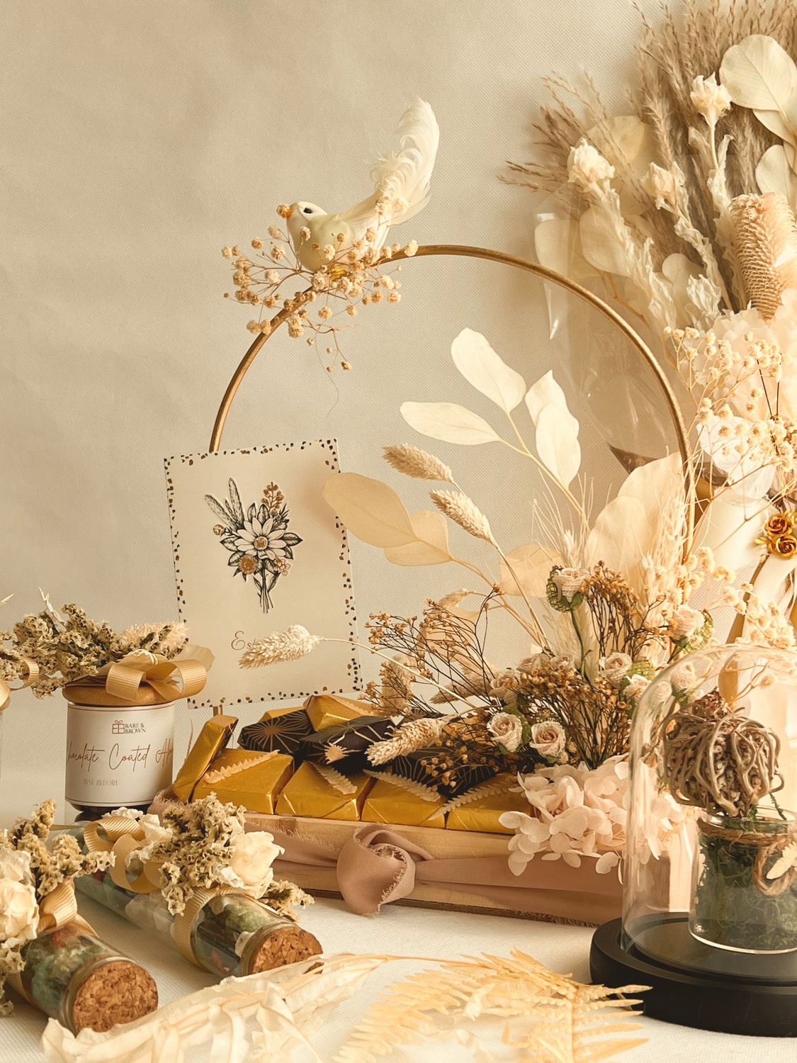 A Bare & Brown composition of dried florals, a gold hoop, branded candle and gold-wrapped confections in warm neutral tones